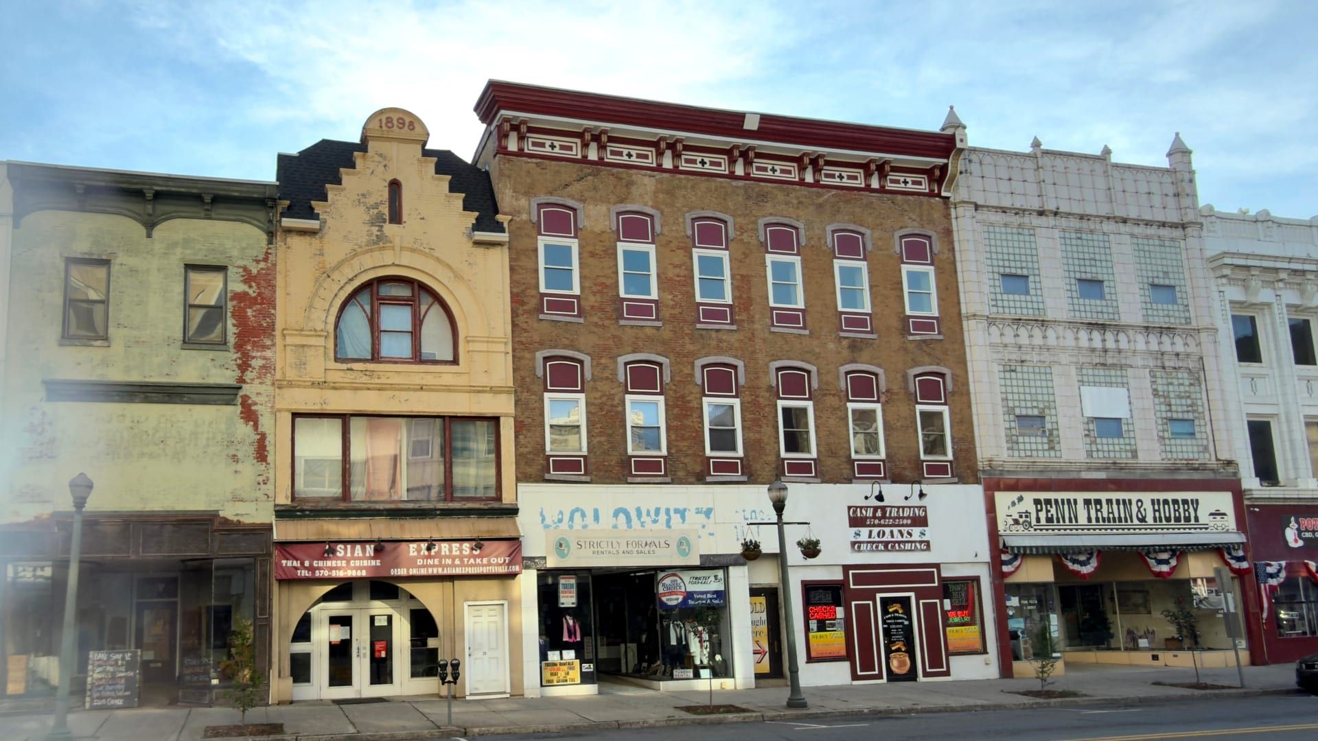 A row of historic storefronts on North Centre Street in downtown Pottsville — small businesses at street level beneath brick, terracotta, and painted upper facades, one stepped gable dated 1898, with patriotic bunting on the shopfronts.