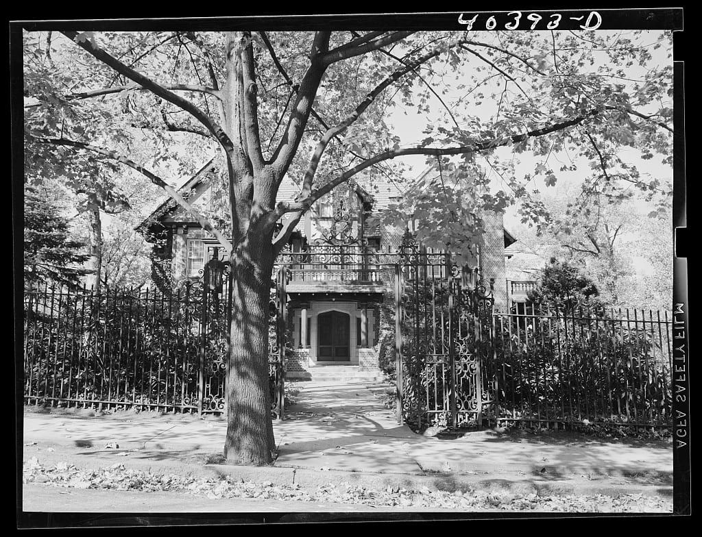 A 1938 photograph of the Frank D. Yuengling mansion in Pottsville: a brick Victorian house with a balcony over its entrance, seen through the bars of an ornate wrought-iron fence with a large tree on the sidewalk.