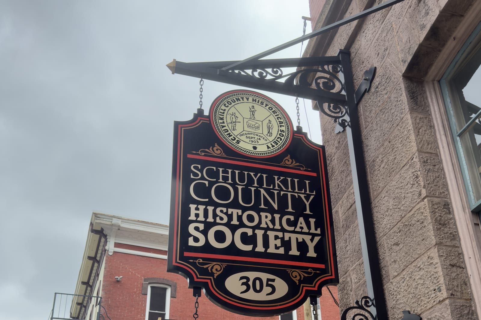 An ornate hanging sign lettered 'SCHUYLKILL COUNTY HISTORICAL SOCIETY / 305' with the society's seal, mounted on a wrought-iron bracket on the stone building at 305 North Centre Street, Pottsville.