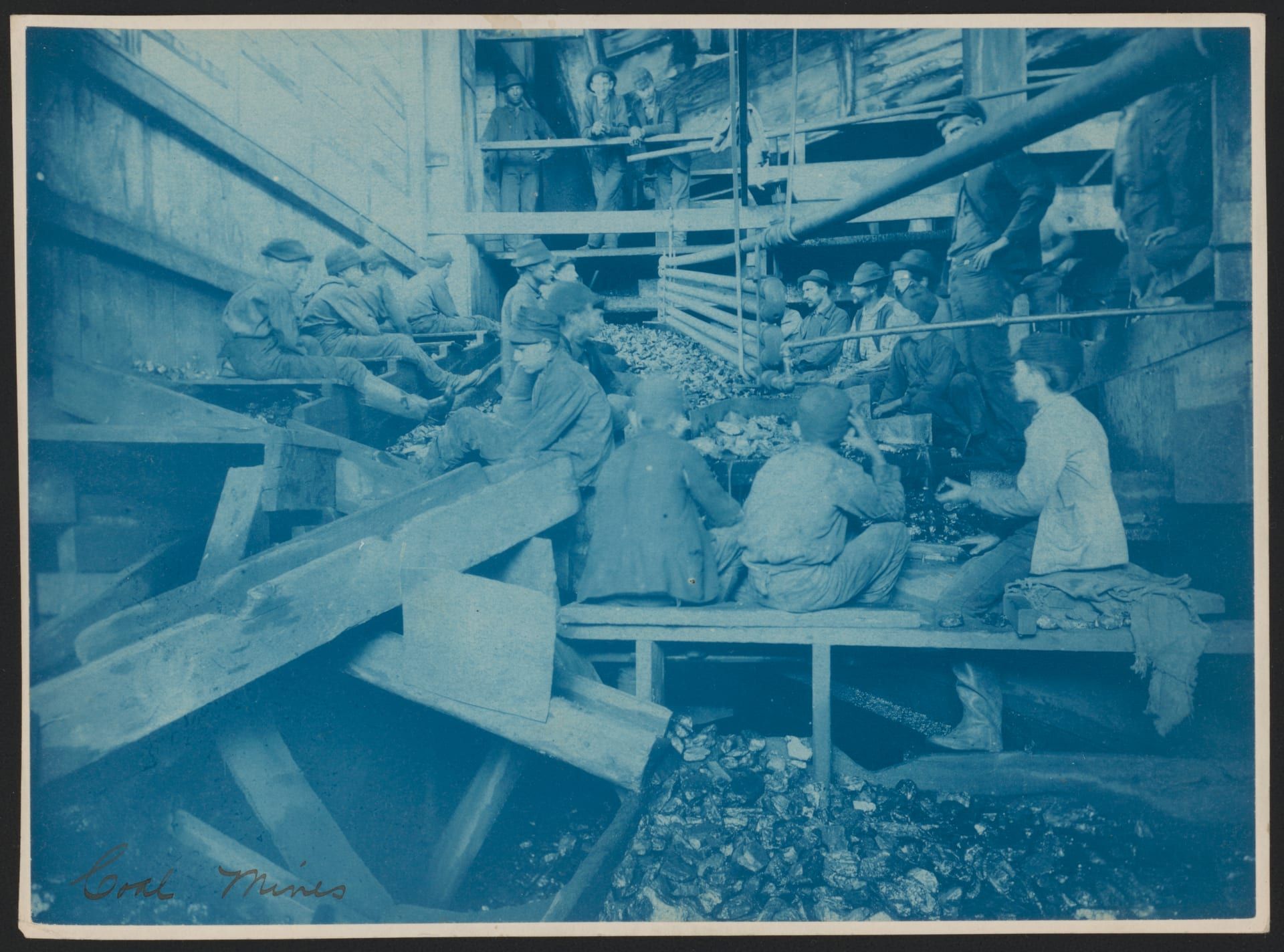 A blue-toned 1891 photograph inside a coal breaker: rows of breaker boys and men perched on the wooden picking chutes, sorting slate from the coal by hand as it passes down the troughs.