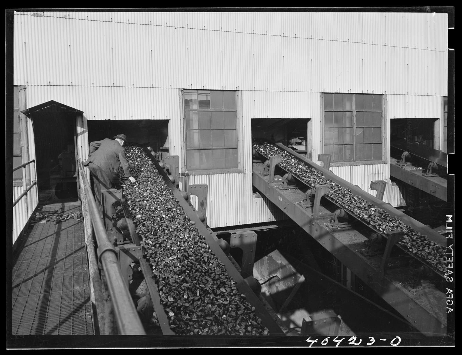 A 1938 interior view at the Saint Nicholas breaker: a company inspector in a suit and hat leans over a steeply inclined belt heaped with broken anthracite, one of several parallel conveyors running past a windowed corrugated-metal wall.