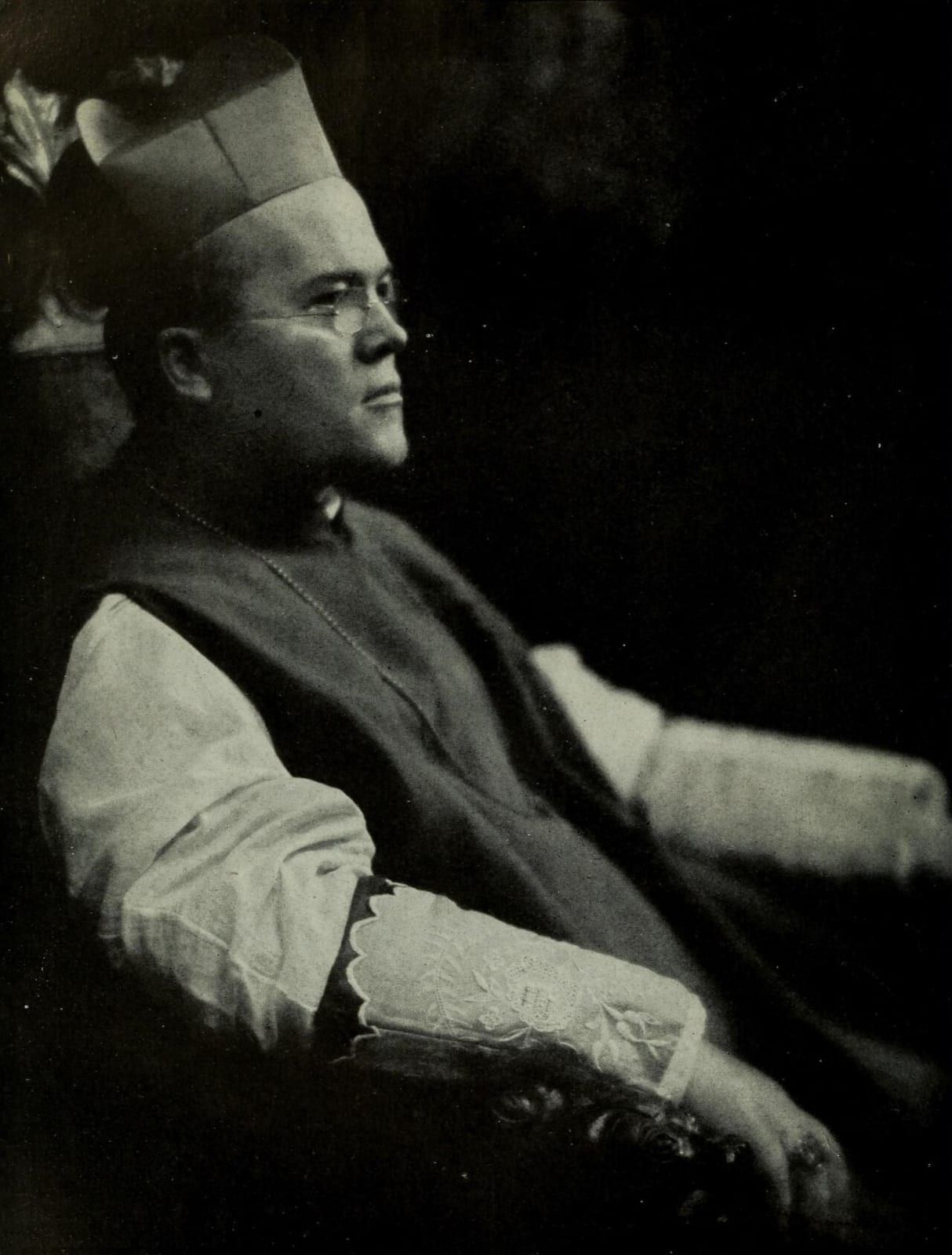 A black-and-white profile portrait of Cardinal Dennis Joseph Dougherty, seated in a biretta, lace-trimmed rochet, and cape with a pectoral-cross chain, wearing pince-nez glasses.