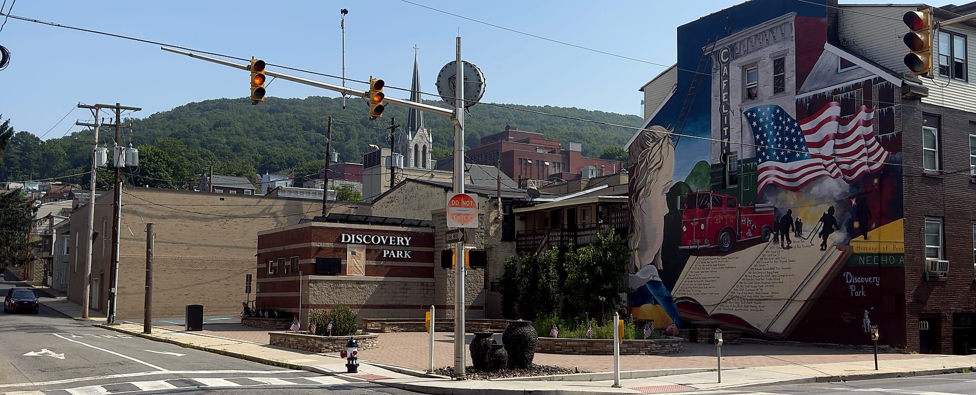 Discovery Park at the corner of Third and West Market streets in downtown Pottsville — a brick plaza with American flags and a painted fire hydrant beneath the DISCOVERY PARK sign, Megan Price's firefighting-history mural on the wall to the right, and a church steeple and wooded ridge rising behind.