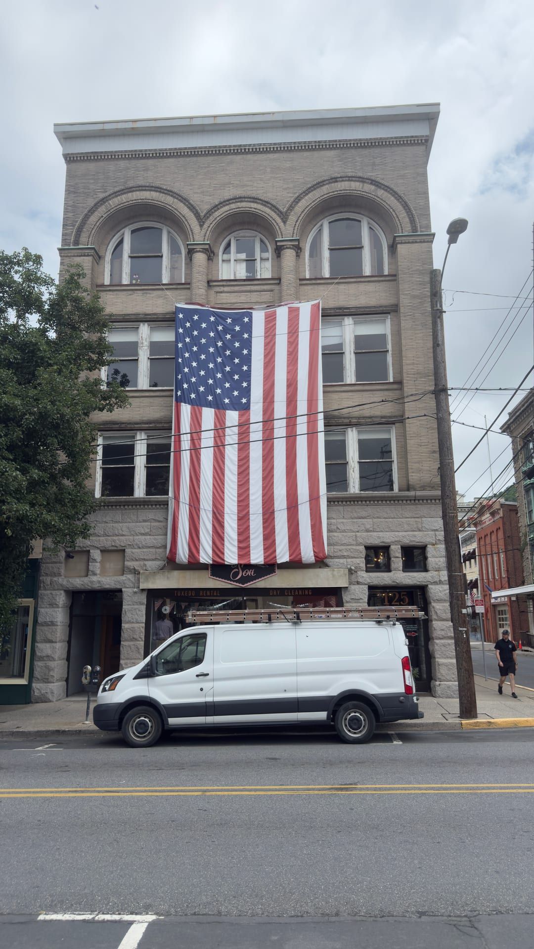 The Hasler Building at 123–125 West Market Street, Pottsville — a four-story commercial block with a rock-faced stone ground floor, three brick upper stories, and a top floor of three round-arched windows, a large American flag draped down the front above the Drasdis & Son storefront (its windows lettered 'Tuxedo Rental' and 'Dry Cleaning').