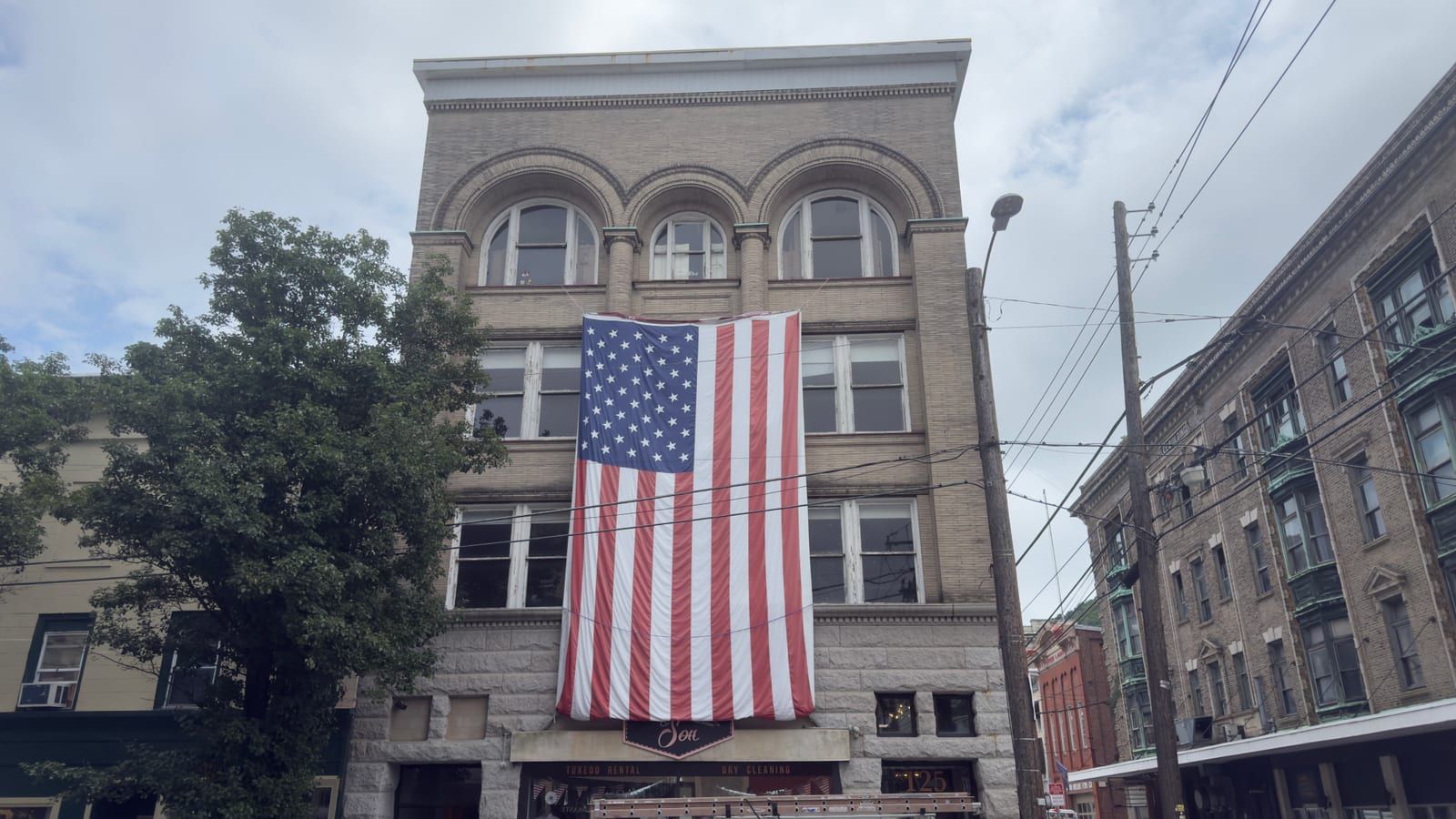 The Hasler Building on the south-east corner of Second and West Market streets in downtown Pottsville — its four-story brick front and draped American flag flanked by neighboring downtown storefronts and commercial blocks, with a large tree at left and utility poles overhead.