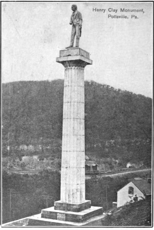 A c.1910 halftone photograph captioned 'Henry Clay Monument, Pottsville, Pa.': the fluted stone column and statue of Clay rising on its stepped base above a wooded hillside, houses and a rail yard below.