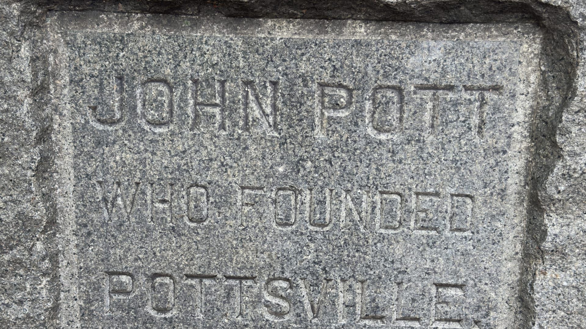 A close view of the John Pott Memorial in downtown Pottsville — deeply carved letters in a rough-hewn granite shaft reading 'JOHN POTT / WHO FOUNDED / POTTSVILLE.'