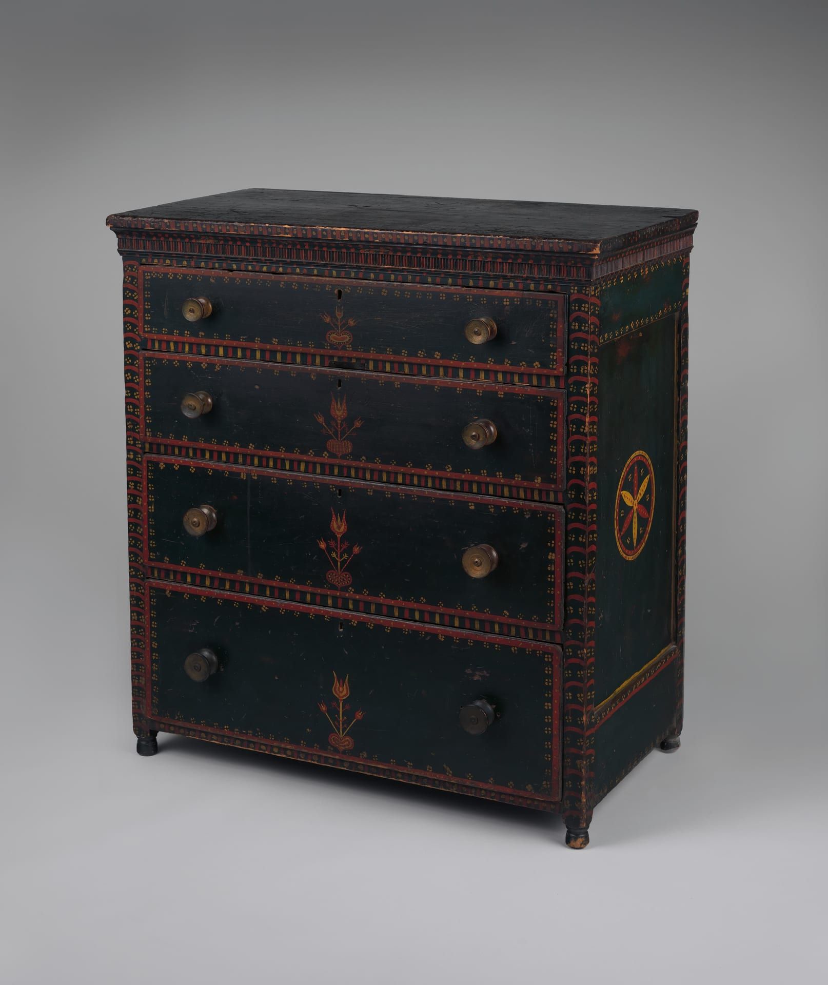 A four-drawer Pennsylvania German chest of drawers painted deep green, each drawer centered with a red-and-yellow tulip sprig rising from a heart, bordered by rows of tiny yellow dots and red-and-yellow banding, with turned wooden knobs and a red-and-yellow pinwheel roundel on the side panel.