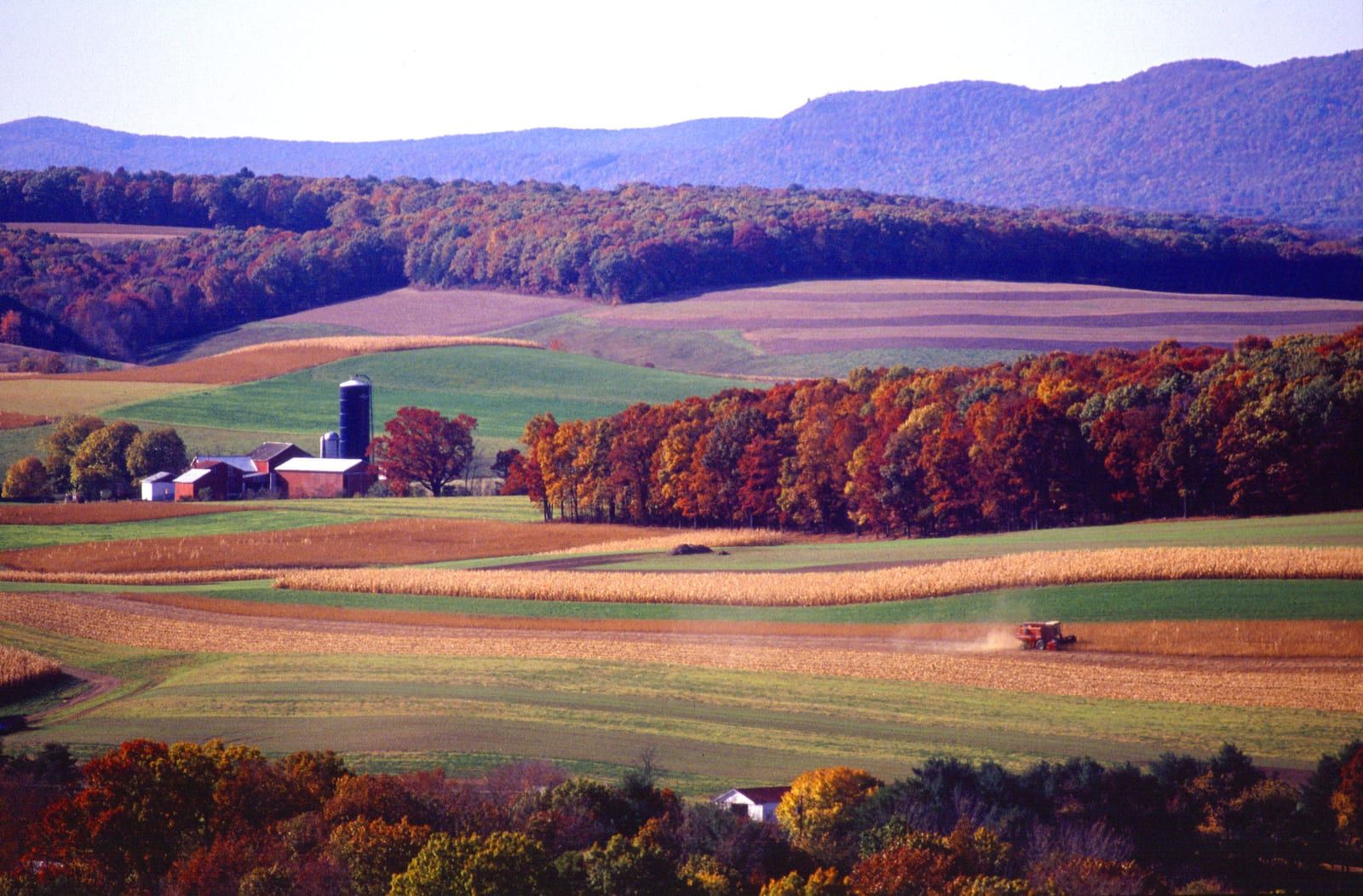 Rolling contour-striped farm fields in green and gold at harvest time, a combine raising dust in a cornfield, a red farm with two silos among brilliant autumn woodlots, and a long forested ridge closing the valley behind.