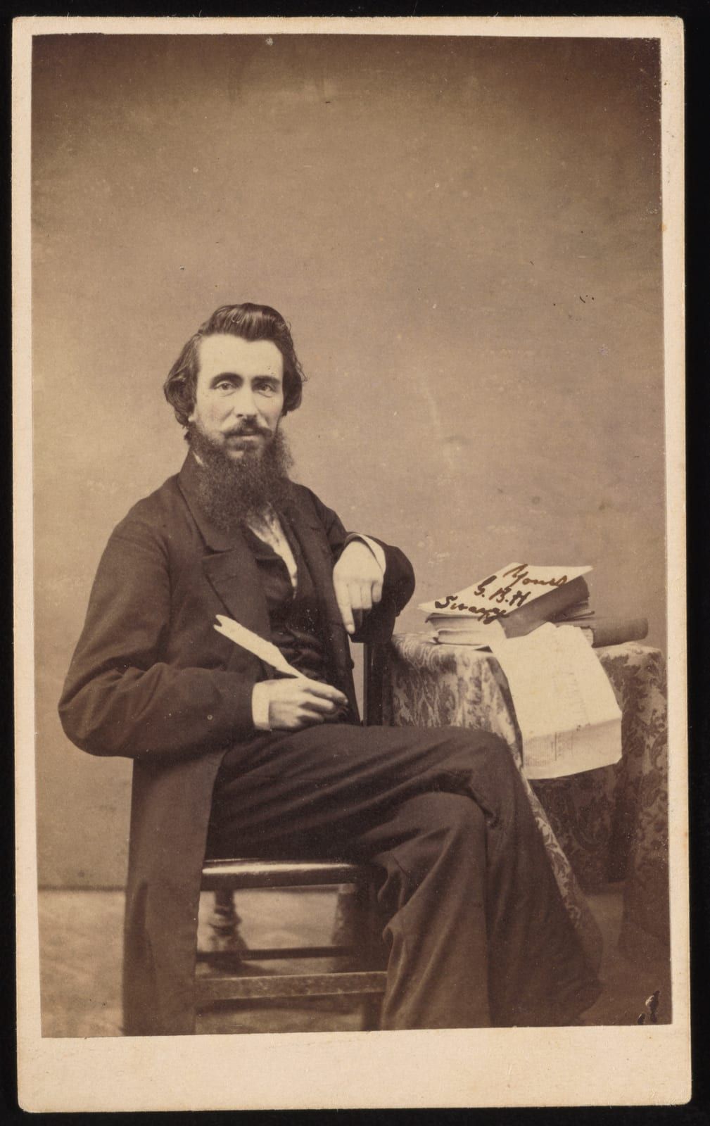 A carte de visite of a seated young man with a full dark beard, in a dark frock coat, holding a quill pen, one arm on the chair back, beside a cloth-draped table stacked with books and papers — a slip on the table signed 'Yours, G.B.H. Swayze.'