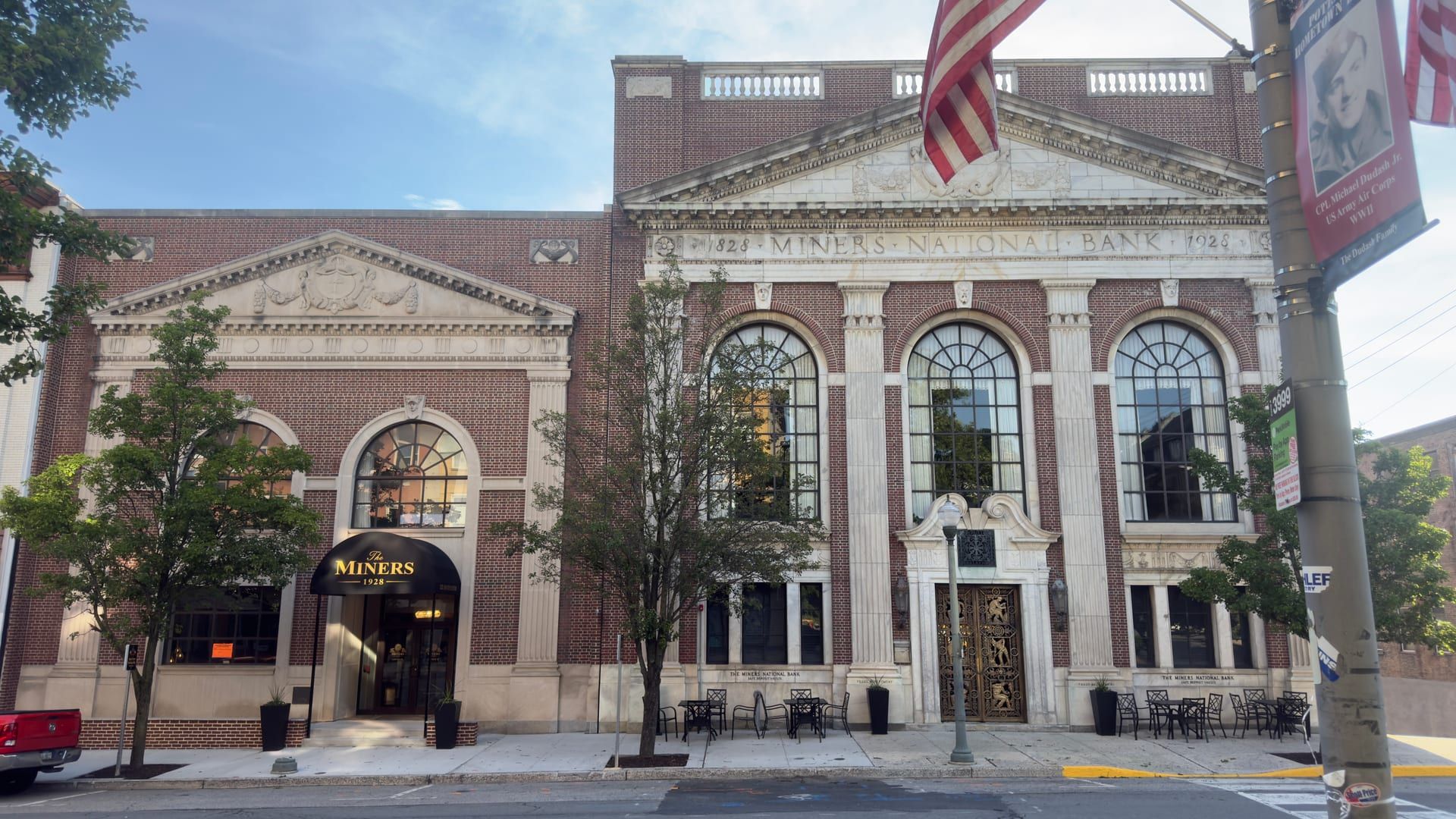 The Miners National Bank building at 120 South Centre Street in Pottsville, now the hotel The Miners 1928 — its central marble pavilion of three tall arched windows between pilasters, beneath a carved pediment and the entablature 'Miners National Bank 1828 1928,' with bronze mining-scene doors at the entrance and the hotel's awning at the adjoining bay.