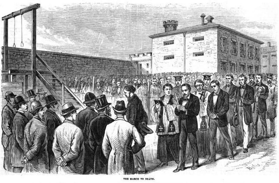 An 1877 wood engraving, 'The March to Death': condemned Molly Maguire prisoners led in a line across a prison yard past a wooden gallows, accompanied by clergy and watched by a crowd, the prison building behind.