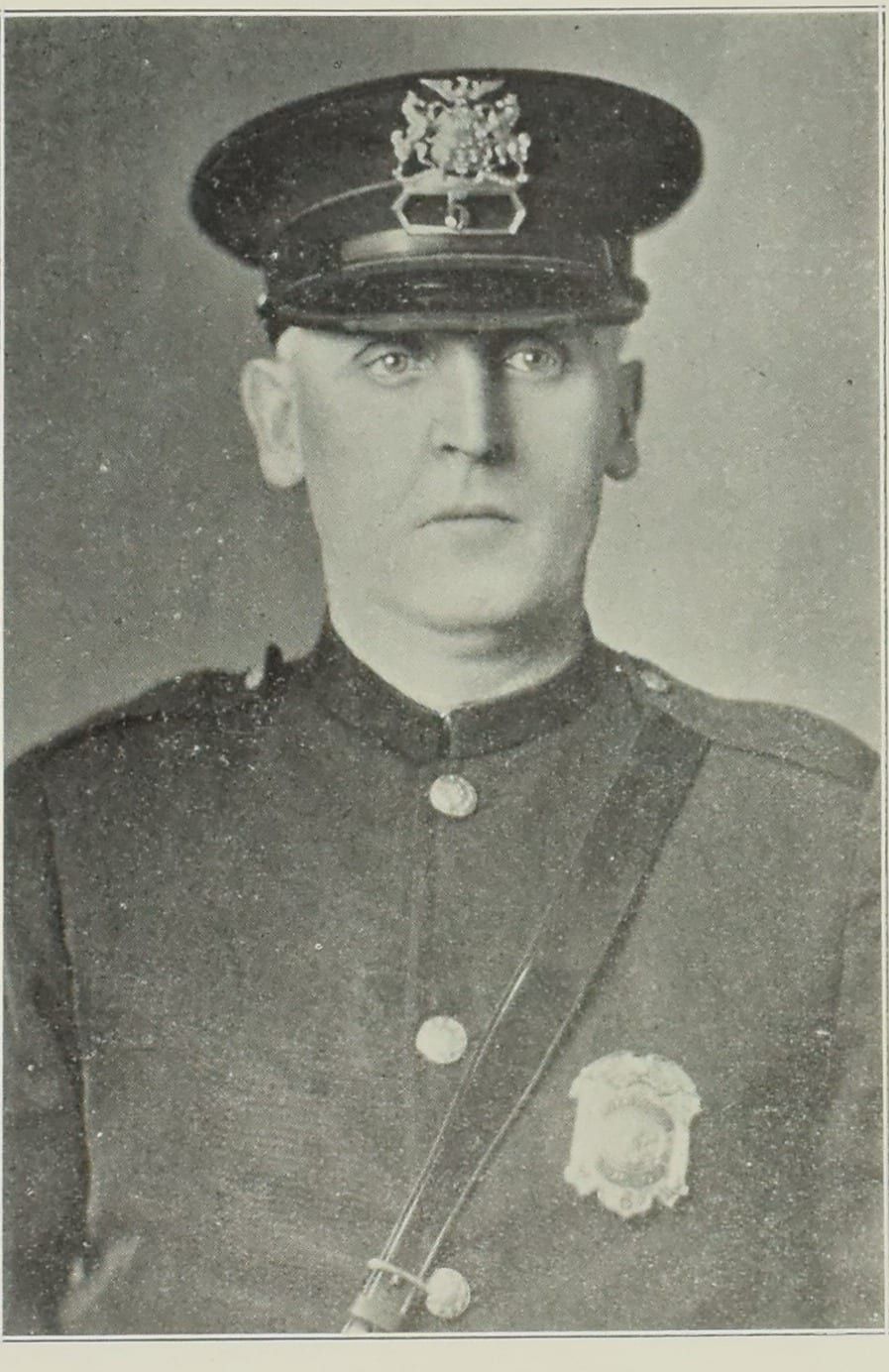 A 1927 halftone photograph of Patrolman Morgan E. Skidmore, a broad-faced middle-aged officer looking straight at the camera in a peaked cap with an eagle-crested badge plate and a high-collared dark tunic with a shield badge on the left breast and a leather cross-strap.