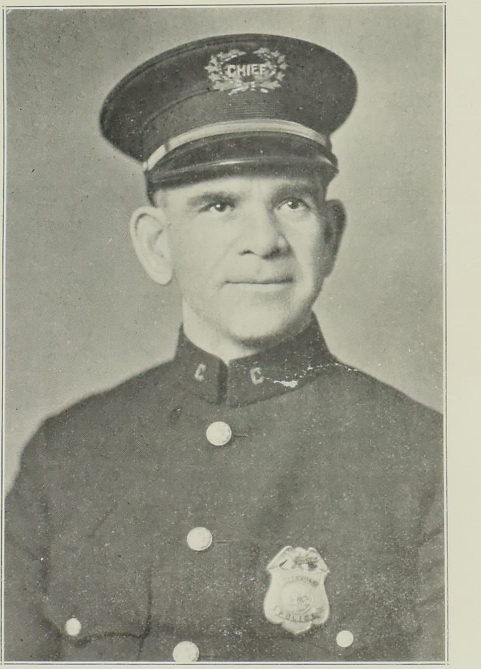 A 1927 halftone photograph of Chief of Police Fred M. Dewald, a trim-faced man with a slight smile in a flat-topped uniform cap whose band reads CHIEF in a wreath, a dark tunic with a C on each collar point, and a shield badge on the left breast.