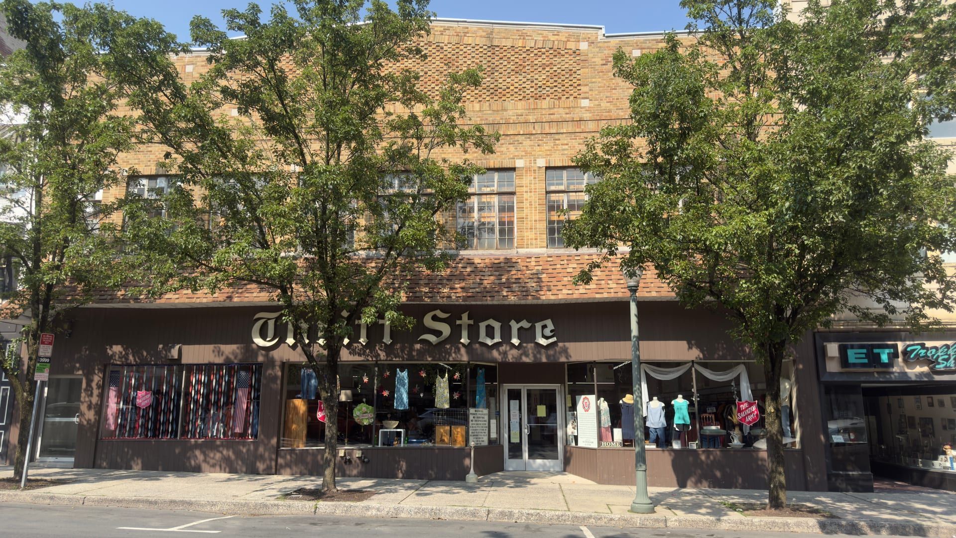 Two-story tan-brick storefront of The Salvation Army thrift store on North Centre Street in Pottsville, its display windows holding clothing and red Salvation Army shield signs beneath a row of street trees.