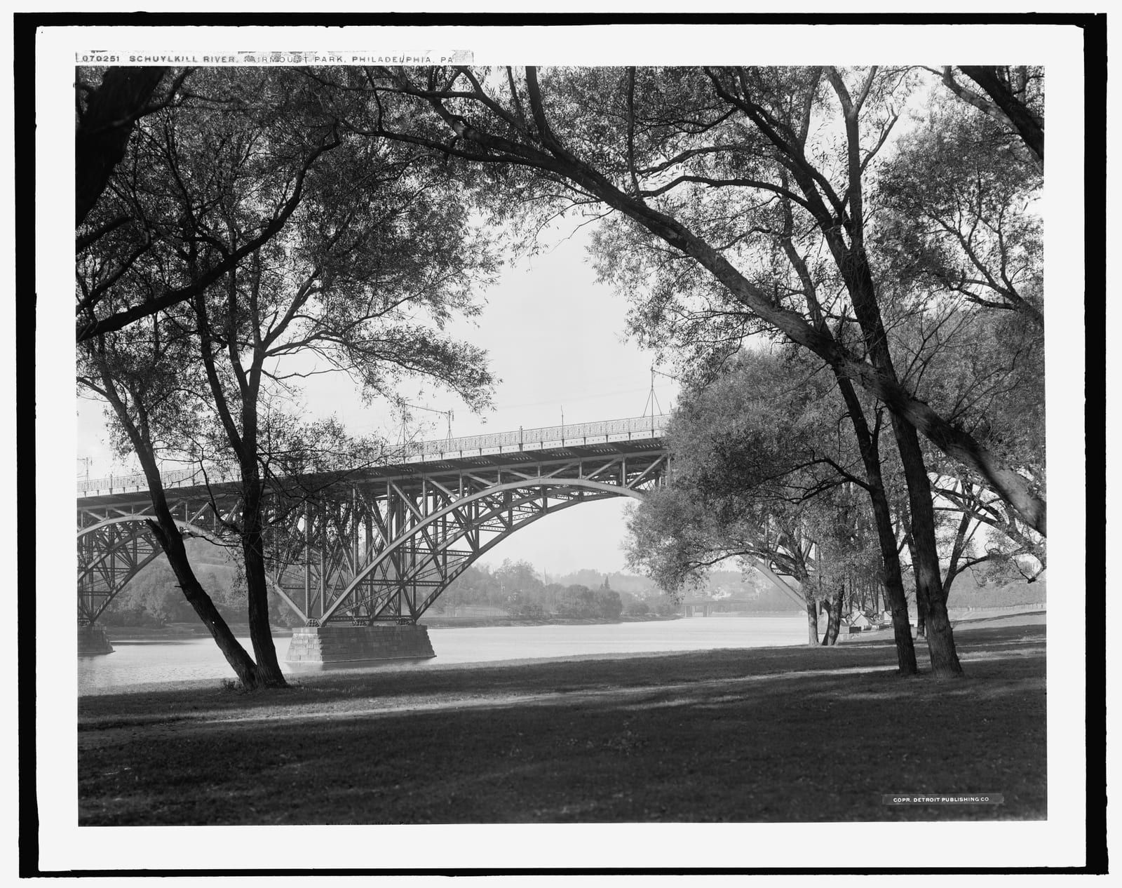 A c.1908 photograph of the Schuylkill River at Fairmount Park, Philadelphia: the steel arch of the Strawberry Mansion Bridge spanning the water, framed by tall willows along a grassy bank downstream.