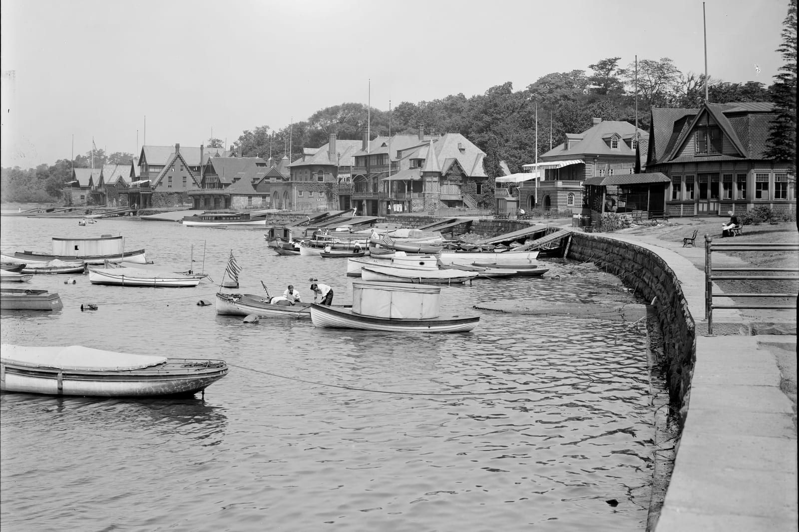 Rowing-club boathouses along the Schuylkill River at Boathouse Row in Fairmount Park, Philadelphia — dozens of moored launches and rowboats, a small boat flying an American flag, and the curved stone river wall and promenade at right.