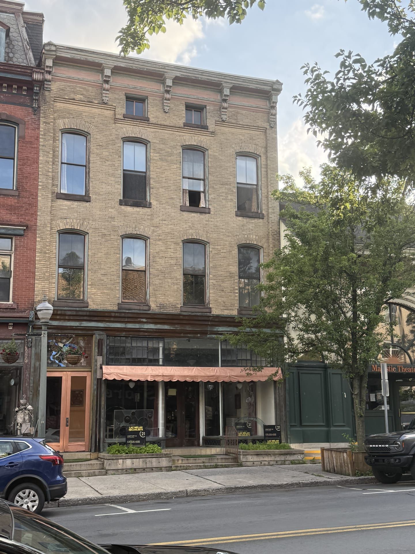 The Ulmer Building at 207 North Centre Street, Pottsville — a three-story buff-brick commercial building with segmental-arched windows and a low attic story under a bracketed cornice; a peach awning shades the vacant storefront, where Century 21 for-sale signs stand in the windows beside the Majestic Theatre.