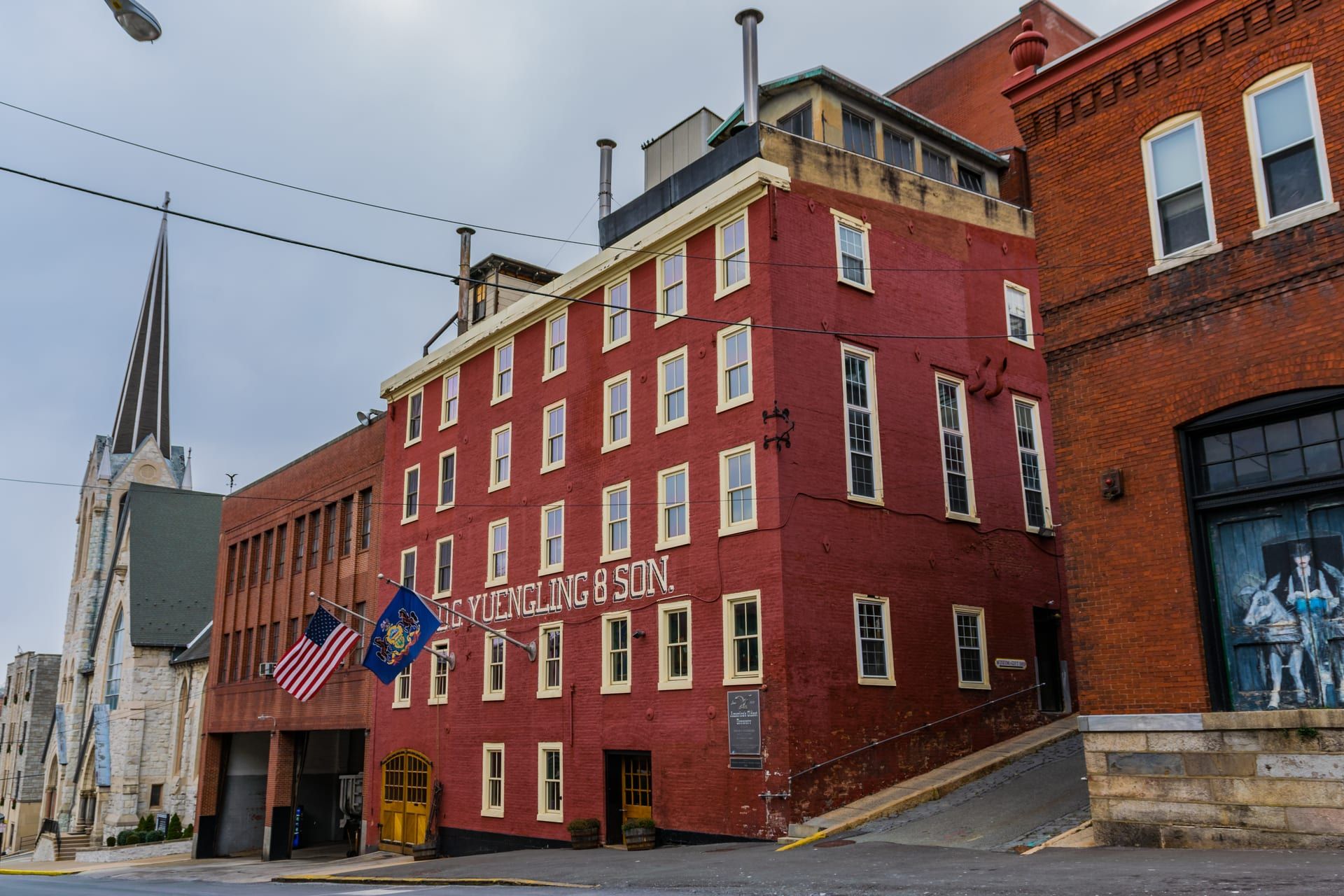 The red-brick D.G. Yuengling & Son brewery on Mahantongo Street in Pottsville, its name painted across the upper facade, American and Pennsylvania flags projecting over the entrance, a church steeple rising beside it.