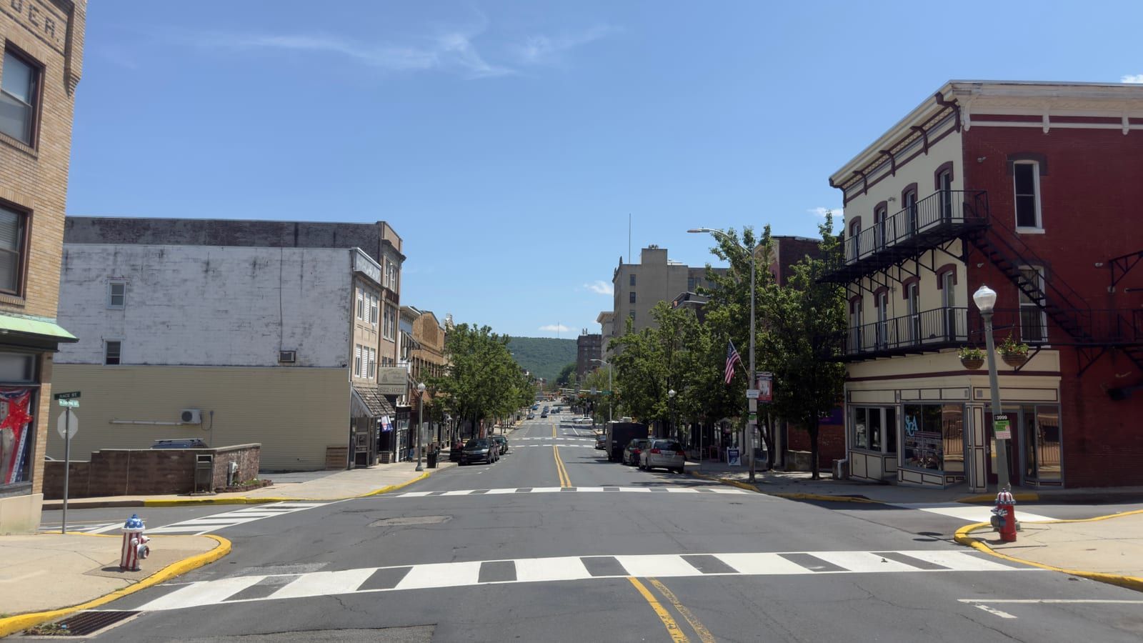 A view down North Centre Street in downtown Pottsville — an empty two-lane street with a crosswalk in the foreground, lined with two- and three-story brick commercial buildings including a red-brick block with iron balconies at right, closing on a wooded ridge in the distance.