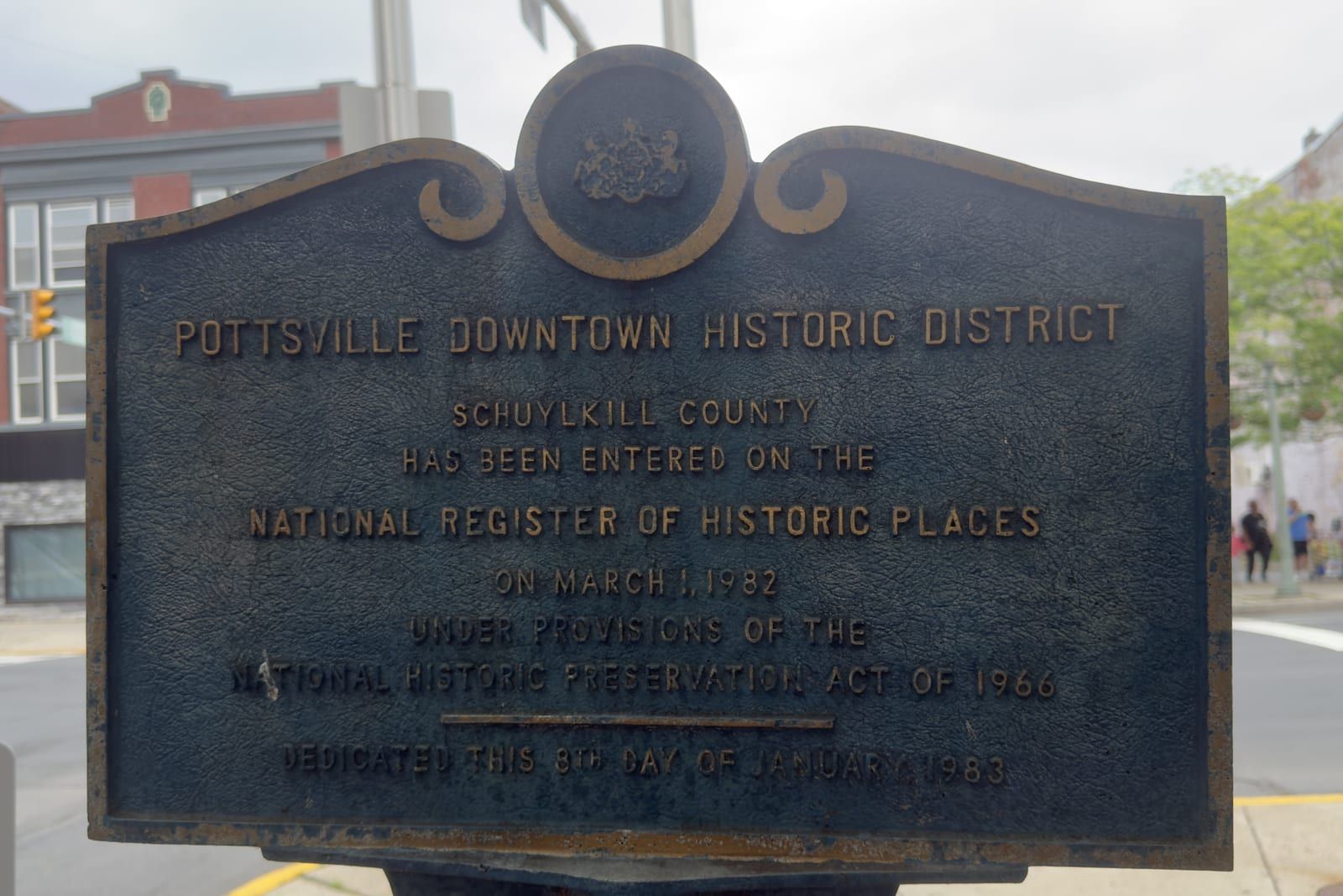 A cast bronze plaque reading 'POTTSVILLE DOWNTOWN HISTORIC DISTRICT — Schuylkill County has been entered on the National Register of Historic Places on March 1, 1982,' mounted at a downtown Pottsville street corner.